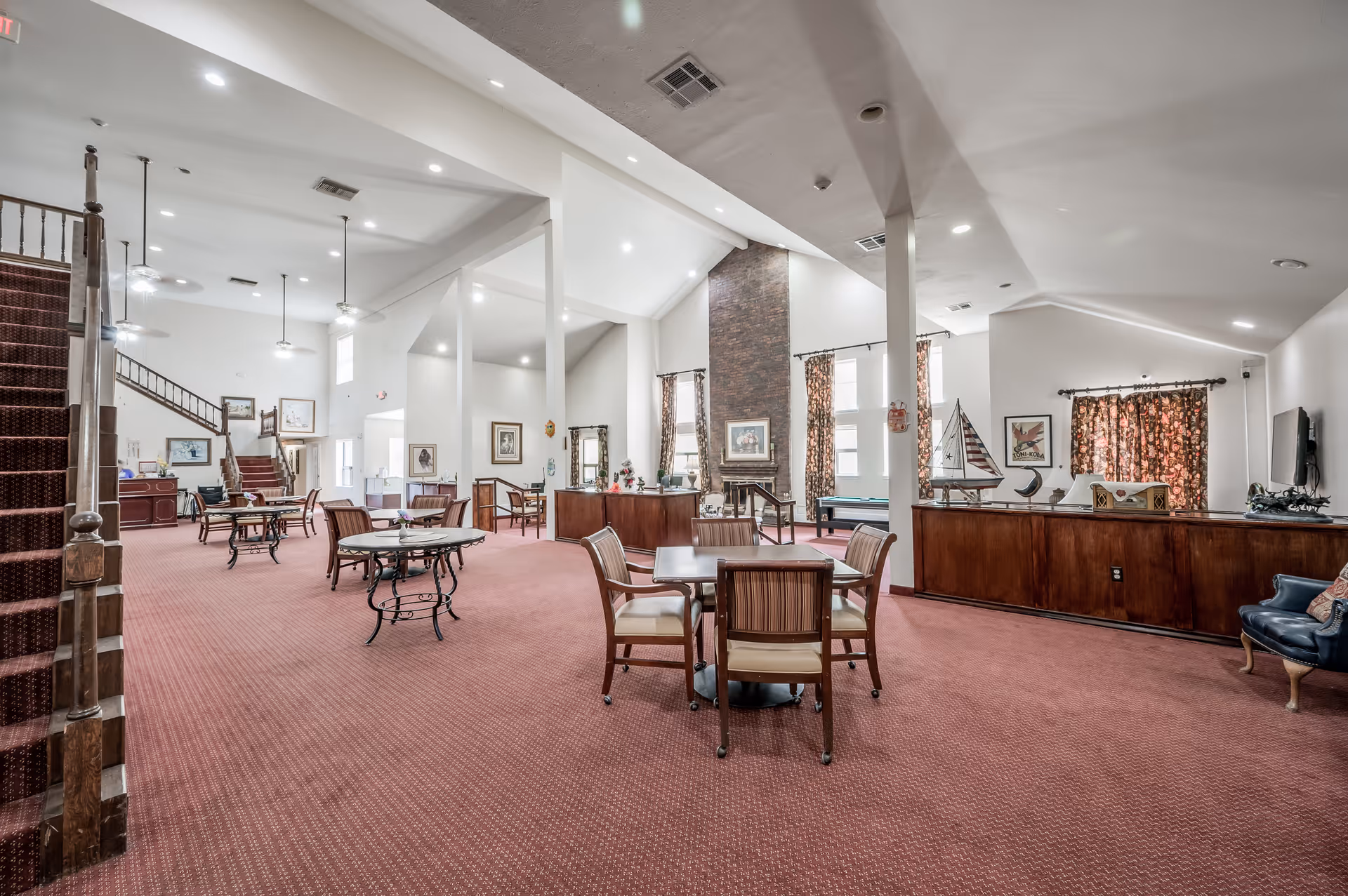 Spacious senior living common area with red carpet flooring, multiple tables and chairs, a staircase on the left, a brick fireplace in the center, and large windows with patterned curtains allowing natural light.