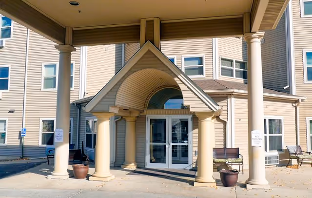 Entrance of a senior living facility with a covered portico supported by large columns. The building exterior is beige with multiple windows, and there are benches and potted plants near the entrance. Signs indicating no parking are visible on the columns.