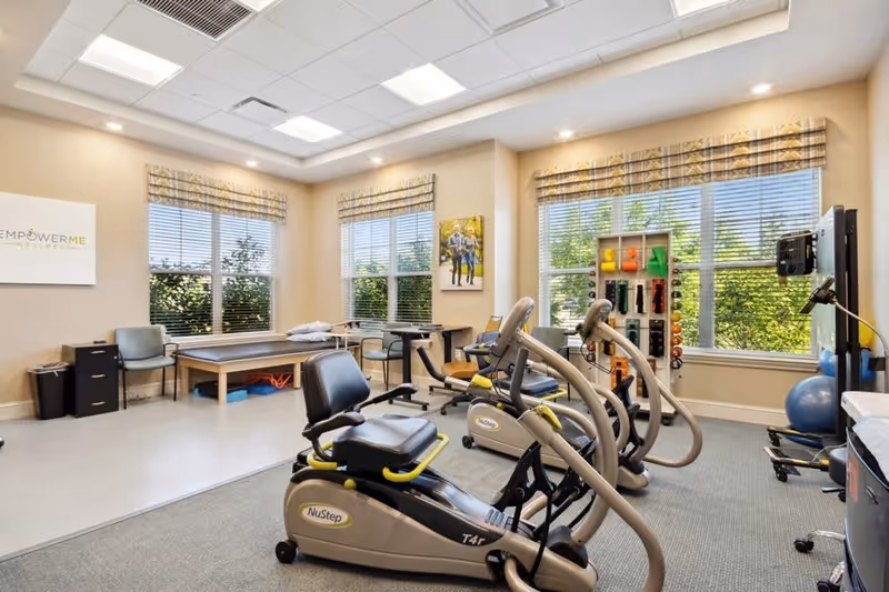 A bright exercise room with large windows letting in natural light. The room contains two NuStep recumbent cross trainers, a rack of colorful hand weights, an exercise ball, and a padded therapy table. There are chairs and a small table near the windows, and the walls are decorated with a sign that reads 'Empower Me' and a framed picture of two people walking outdoors.