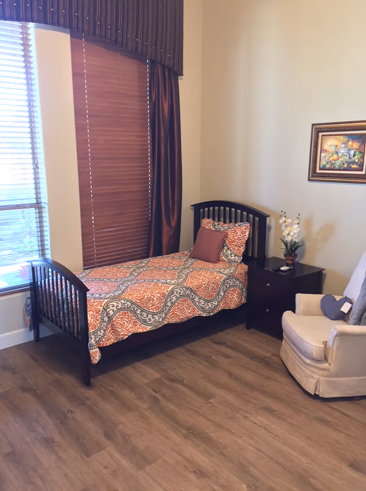 Single bed with patterned orange bedding, a nightstand, armchair, and window with blinds in a senior living bedroom.