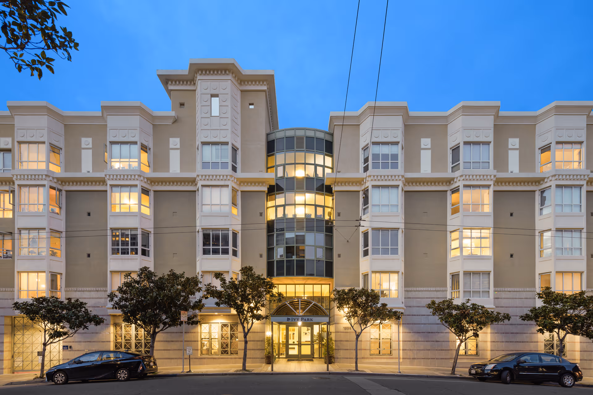 Exterior front view of a multi-story residential building named Ivy Park at Cathedral Hill, with large windows, trees lining the sidewalk, and cars parked on the street in front. The building is illuminated from inside during dusk.