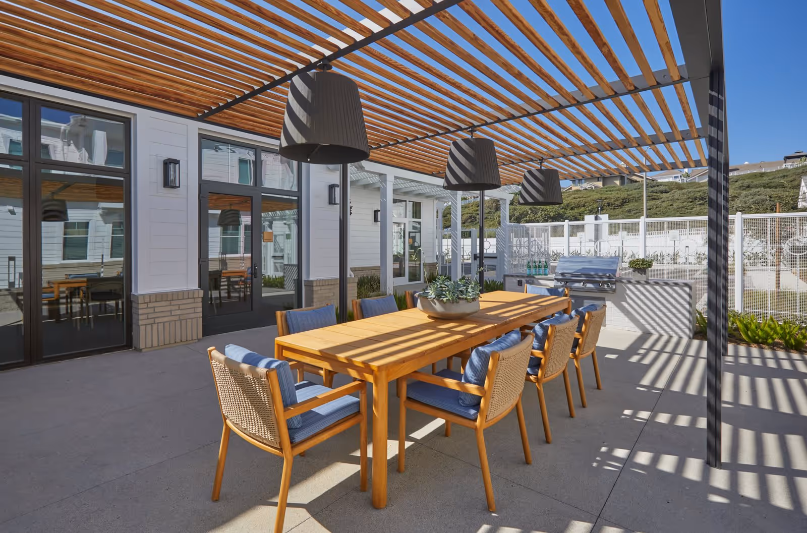Covered outdoor dining patio with a wooden table and chairs under a slatted pergola next to building windows.