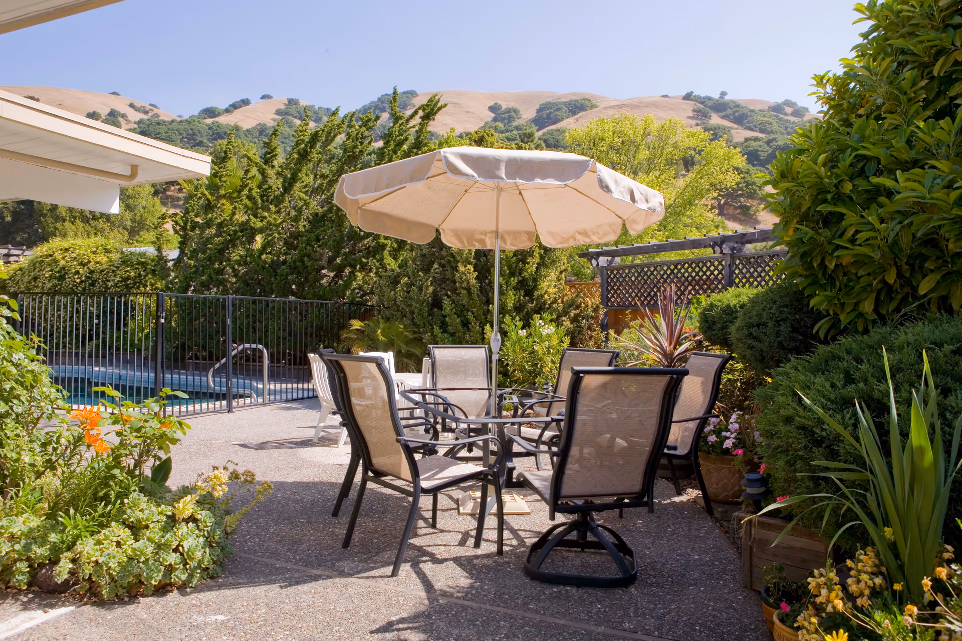 Outdoor patio area with a round table surrounded by six mesh chairs and a large beige umbrella. The patio is surrounded by lush green bushes and plants, with a swimming pool fenced in the background and hills visible under a clear blue sky.