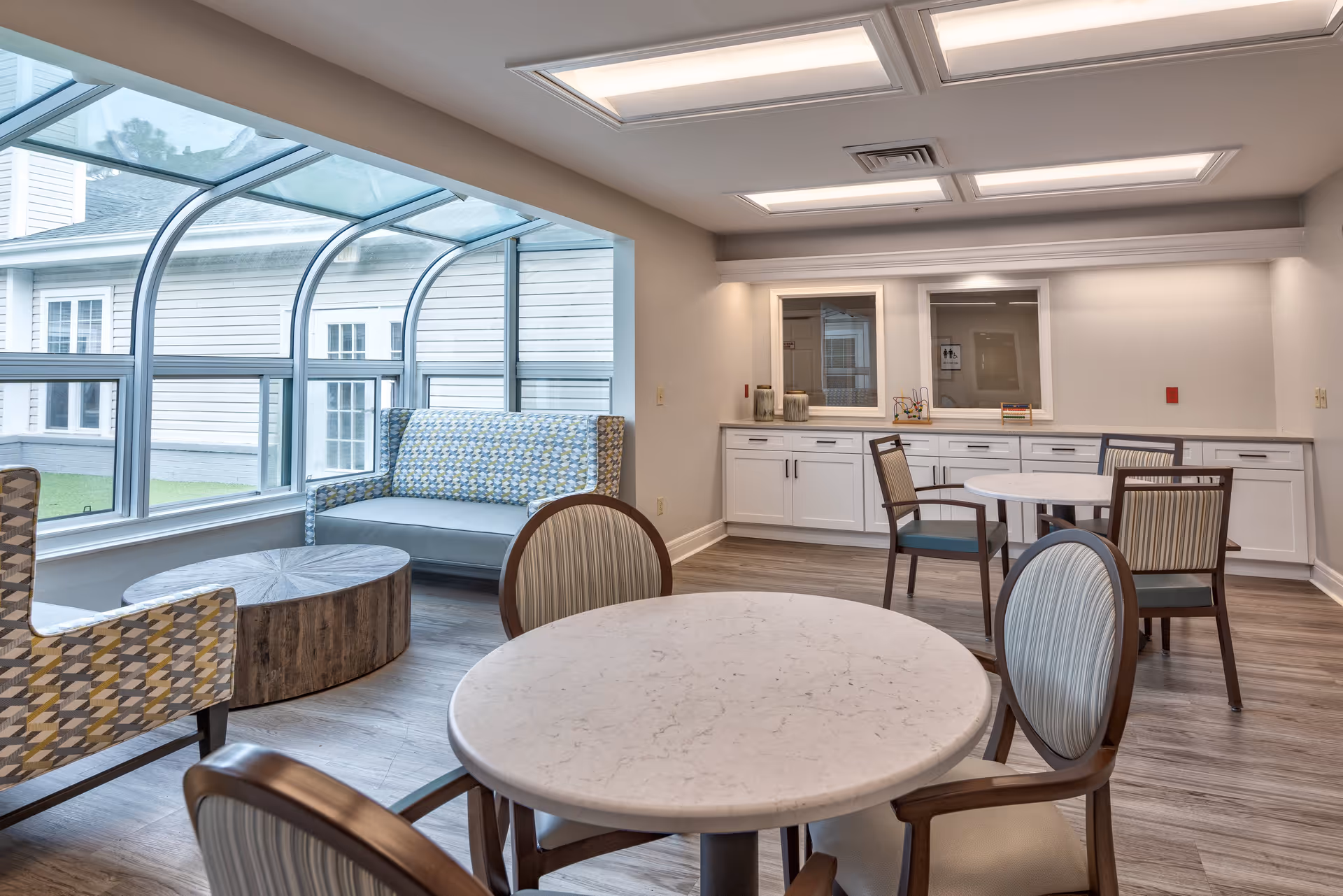 A bright and modern common area with round tables and chairs, a patterned armchair, a patterned loveseat, and a wooden coffee table. Large curved windows provide natural light and a view of the exterior building and grass.