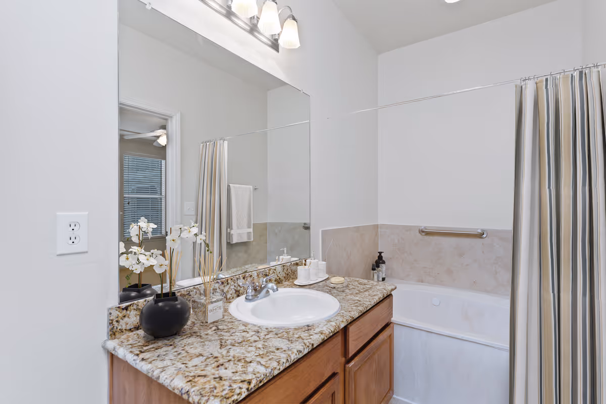 A clean bathroom featuring a granite countertop with a white sink, a large mirror above the sink, a bathtub with a striped shower curtain, and a towel hanging on the wall. Decorative items including a black vase with white flowers and a reed diffuser are placed on the countertop.