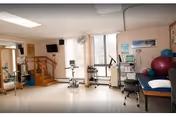 A bright physical therapy or rehabilitation room with exercise equipment including stationary bikes, a treadmill, therapy balls, and a treatment table. The room has large windows letting in natural light, a wall-mounted TV, and a staircase in the background.