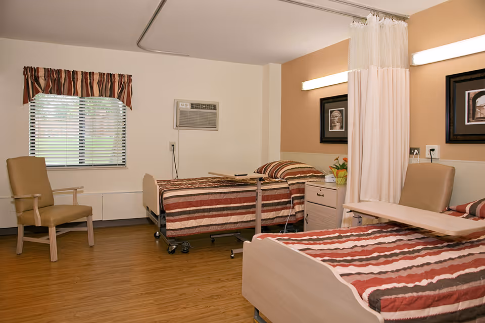 A nursing care center room with two hospital beds, each with striped bedding in shades of brown, red, and beige. There are two beige chairs, a window with blinds and a striped valance, a wall-mounted air conditioning unit, a bedside table with a plant, and a privacy curtain between the beds. The walls are painted beige and white, and the floor has a wood-like finish.