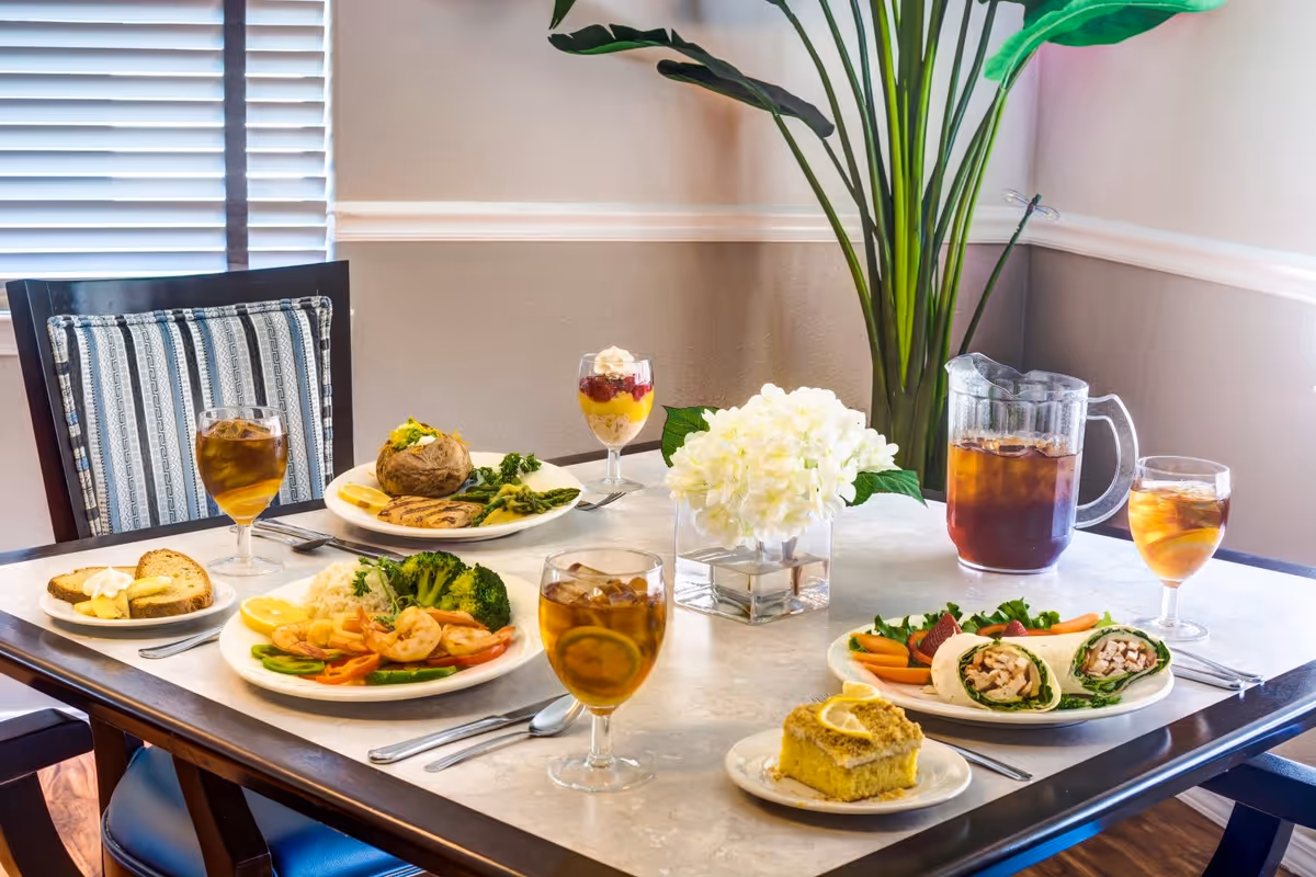 A dining table set with plates of food including shrimp with vegetables, a baked potato with asparagus, a wrap sandwich with carrot sticks, cornbread, and dessert parfaits. There are glasses of iced tea and a pitcher of iced tea on the table. A vase with white flowers and a tall green plant are also on the table. The setting is in a room with a striped chair and window blinds in the background.