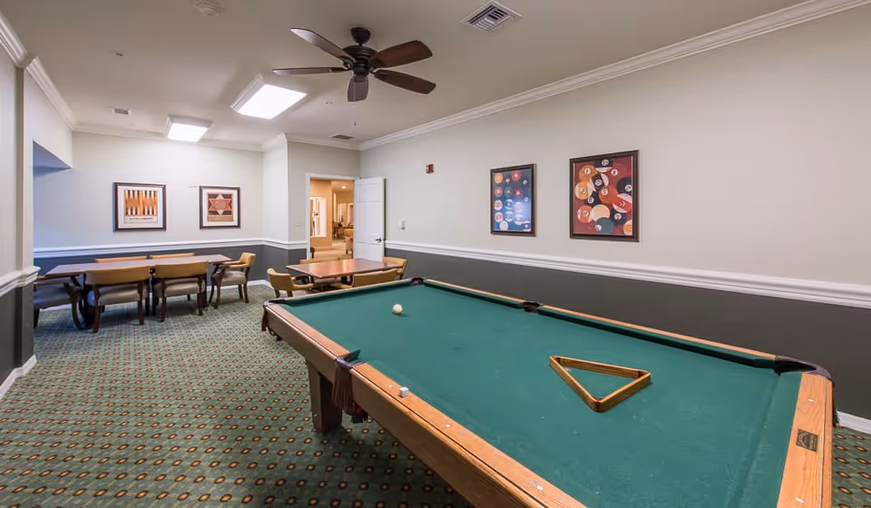 Interior room with a pool table in the foreground and two tables with chairs in the background. The room has patterned carpet, light-colored walls with a dark wainscot, ceiling fan, and framed pictures on the walls.