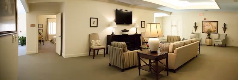 Open seating lounge in a senior living facility with sofas and armchairs arranged around a TV and side tables.