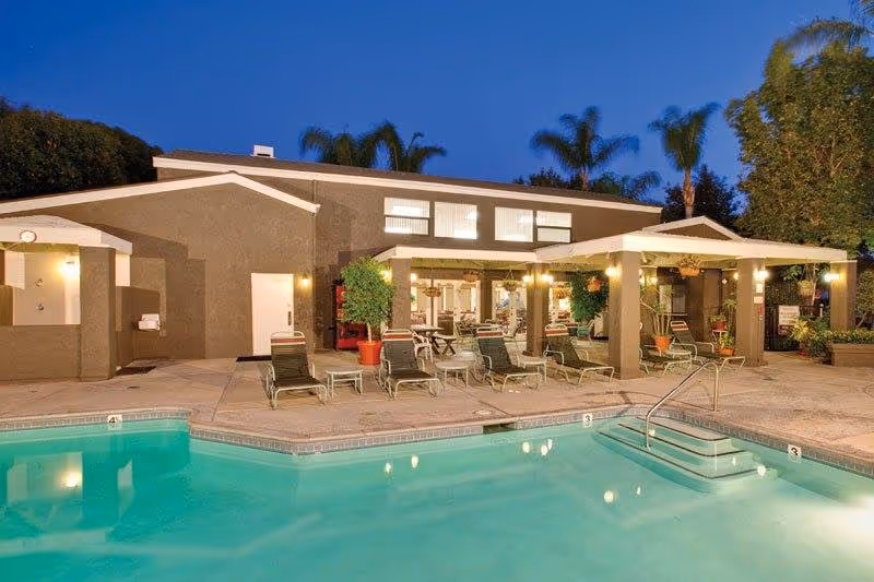 Outdoor swimming pool area at dusk with lounge chairs arranged around the pool. A building with large windows and a covered patio is visible in the background, surrounded by palm trees and other greenery.