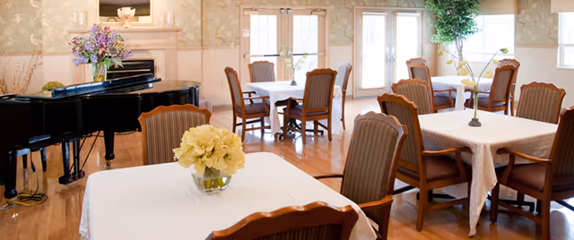 A bright dining room with several tables covered in white tablecloths, each surrounded by wooden chairs with striped cushions. There are flower arrangements on the tables and a black grand piano in the corner. Large windows and glass doors allow natural light to fill the room.