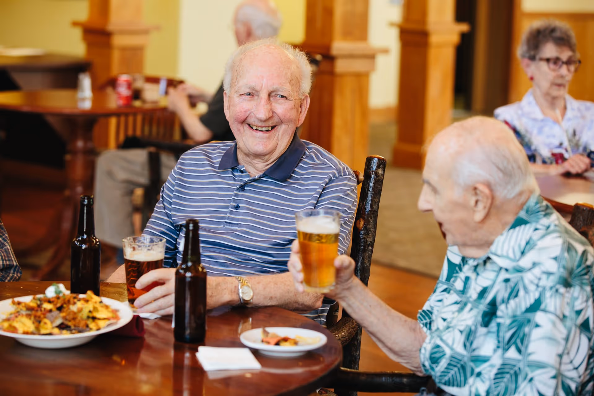 Two elderly men sitting at a wooden table in a communal dining area, smiling and holding glasses of beer. There are plates of food and beer bottles on the table. In the background, other elderly people are seated at tables in a warmly lit room with wooden pillars.