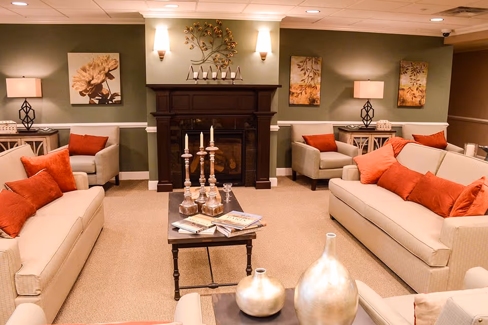 A cozy living room with two beige sofas and two armchairs, all adorned with red and orange cushions. A dark wooden coffee table in the center holds several books and three tall candle holders with white candles. The room features a dark wooden fireplace with decorative wall sconces and artwork above it. Two side tables with lamps and decorative items flank the armchairs. The walls are painted green with white trim, and the floor is carpeted.