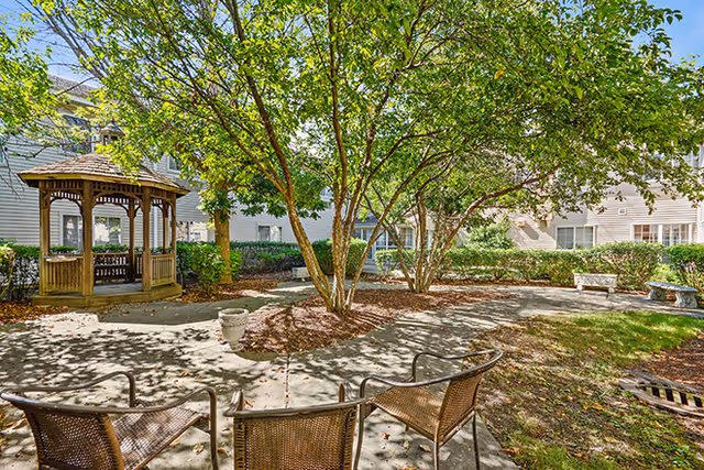 Outdoor courtyard area with a wooden gazebo, several trees providing shade, three metal chairs in the foreground, and stone benches along the pathway surrounded by greenery and residential buildings.