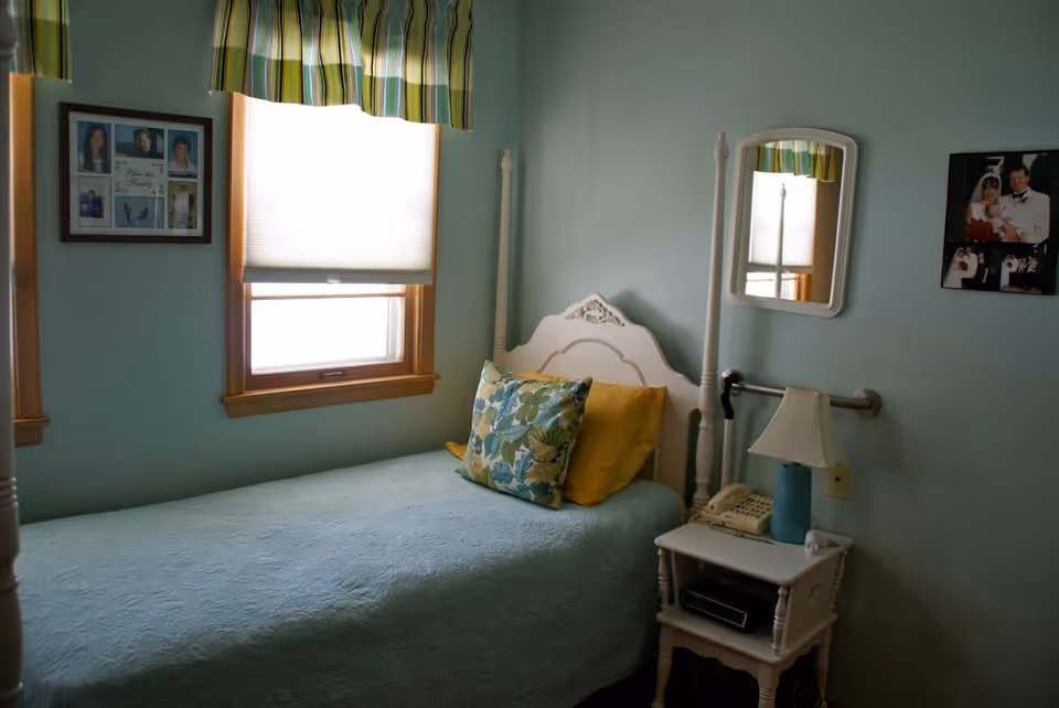 Small single bedroom with a white four-poster bed, mint green walls, patterned pillows, a window with a striped valance, a nightstand with a lamp and phone, and framed photos on the walls.