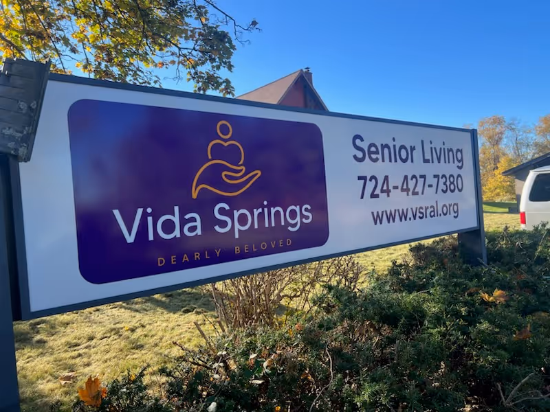 Outdoor sign for Vida Springs Senior Living and Personal Care Home with logo, contact phone number, and website URL, surrounded by greenery and trees under a clear blue sky.