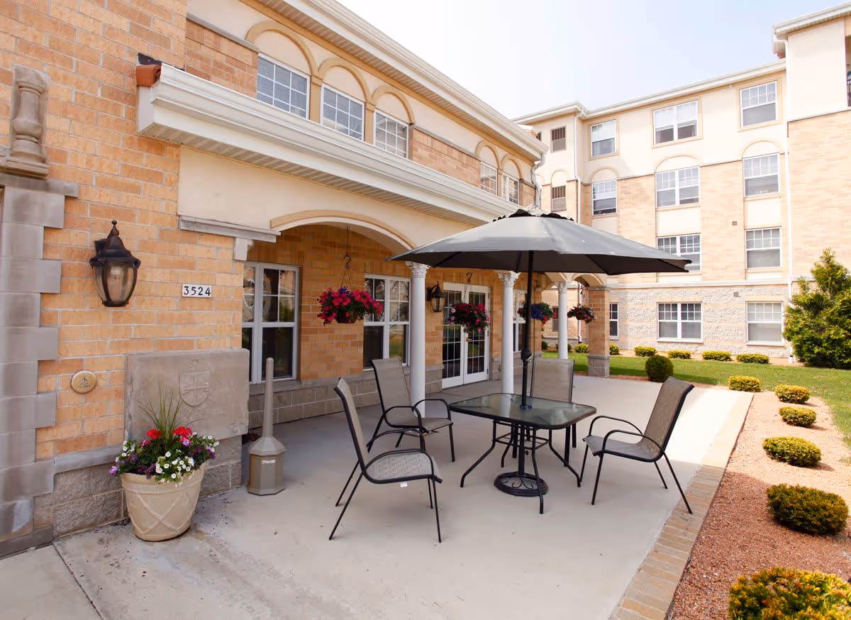 Outdoor patio area at St. Catherine Commons - Independent Living with a glass table, four chairs, and a large umbrella. The patio is adjacent to a brick building with hanging flower baskets and decorative columns. There are neatly trimmed bushes and a small garden area surrounding the patio.