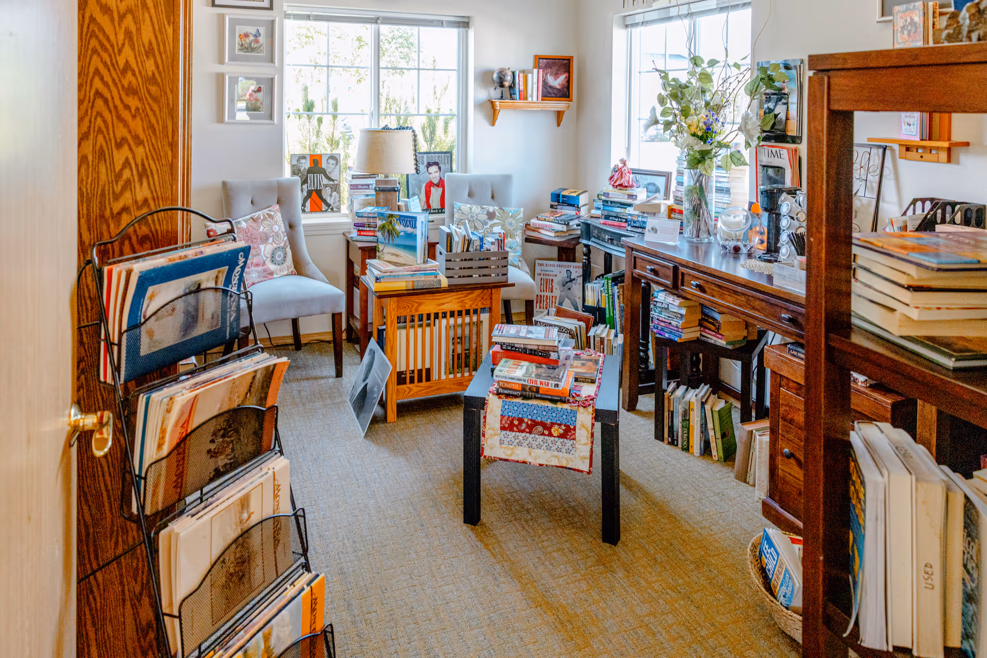 A cozy reading room filled with books on shelves, tables, and racks. Two upholstered chairs with patterned pillows are placed near a window letting in natural light. The room has wooden furniture, including a long console table and a small table with a colorful quilted cloth. Various framed pictures and decorative items are displayed throughout the room.