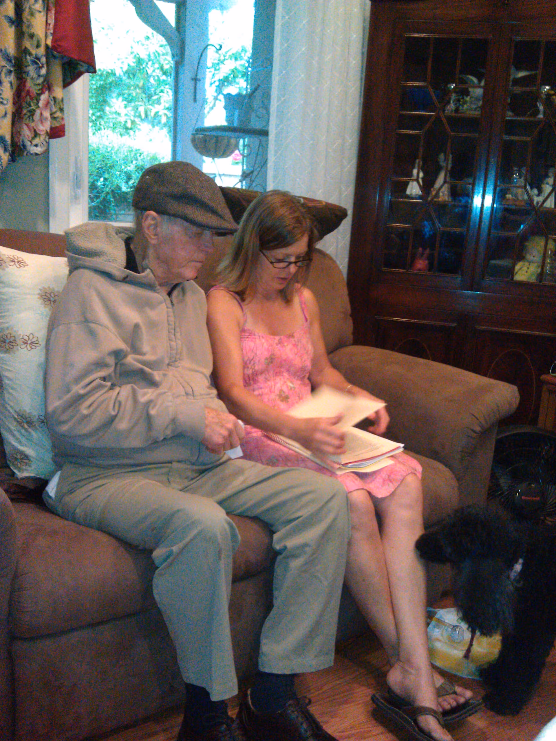 An elderly man wearing a cap and a beige jacket sits on a brown couch next to a woman in a pink dress who is looking at papers. A black poodle stands near the woman's feet. Behind them is a wooden cabinet with glass doors displaying various items, and a window showing greenery outside.