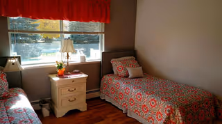 A small bedroom with two single beds covered in matching red and gray floral patterned bedspreads. Between the beds is a white nightstand with two drawers, a table lamp, a small plant, and a book. A window with blinds and a red valance lets in natural light, illuminating the wooden floor and beige walls.