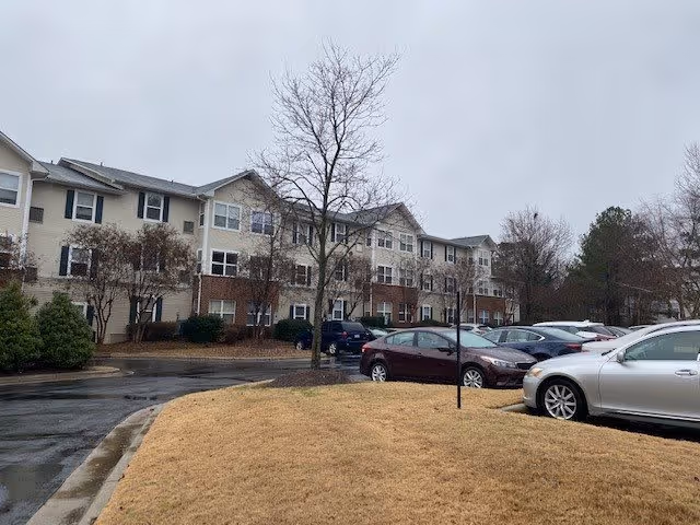 View of a multi-story residential building with beige siding and multiple windows, surrounded by leafless trees and a parking lot with several parked cars on a cloudy day.