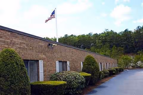 Single-story brick rehabilitation center exterior with shrubs, a driveway, and an American flag.