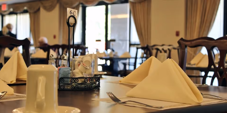 A dining table set with folded beige napkins, silverware, salt and pepper shakers, and a table number holder labeled 6-C in a nursing and rehabilitation center dining room. The background shows more tables and chairs with similar settings and large windows with curtains.