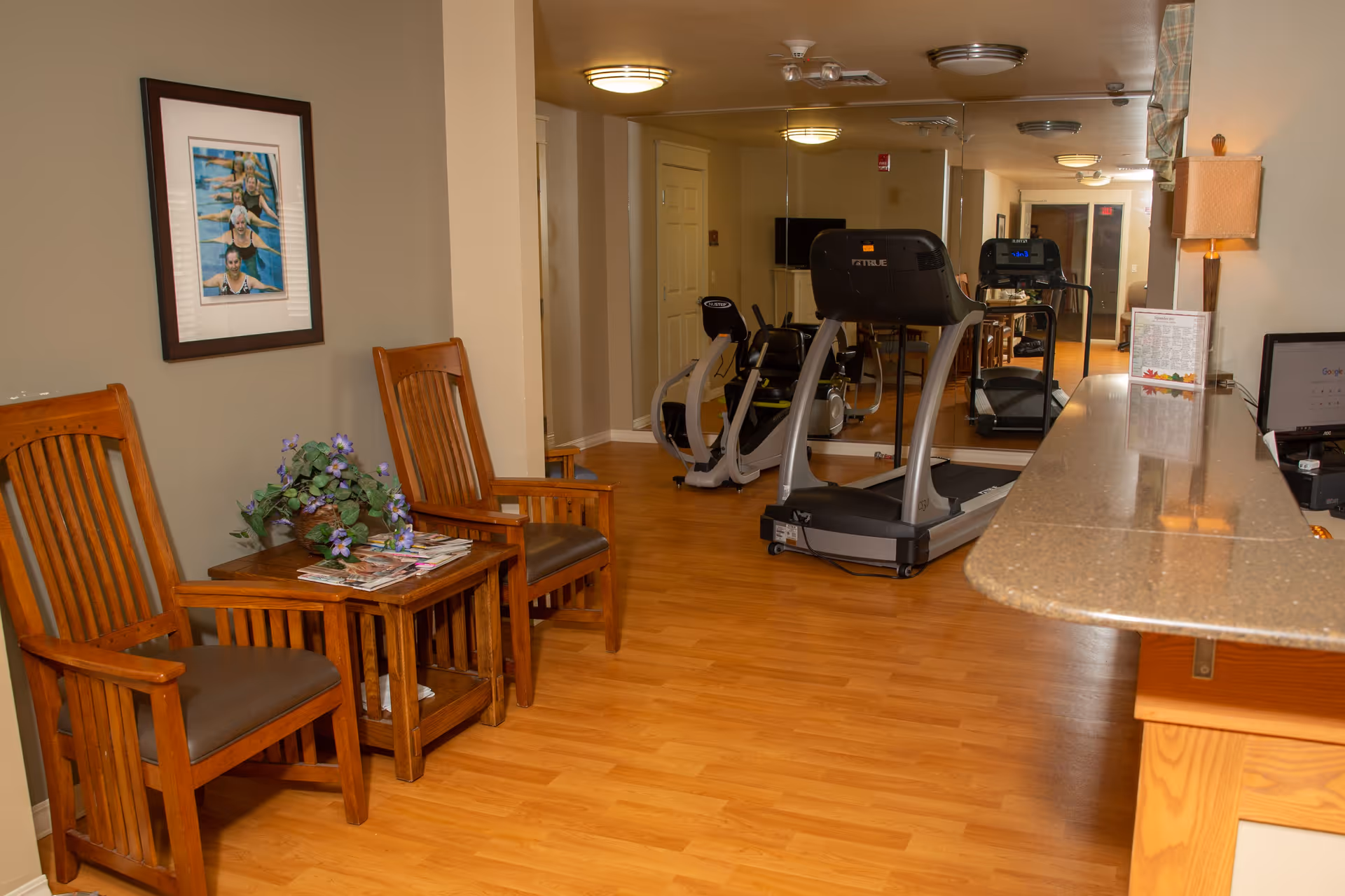 A small fitness area in a senior living facility with a treadmill and a recumbent exercise bike in front of a mirrored wall. To the left, there are two wooden chairs with cushions and a small wooden table with magazines and a potted plant. On the right side, there is a countertop with a lamp and some papers. The floor is wood, and the walls are painted beige with a framed picture hanging above the chairs.