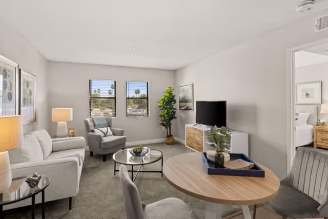 Bright furnished living room with a sofa, armchair, TV on a console, round dining table and decorative plants in a senior living apartment.