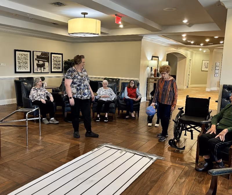 A group of elderly people and a caregiver are gathered in a spacious, well-lit common area with wooden flooring. Some are seated in comfortable chairs arranged in a semi-circle, while one elderly woman is standing and holding a colorful ball. A wheelchair and a walker are visible nearby. The room has neutral-colored walls, framed artwork, and modern lighting fixtures.