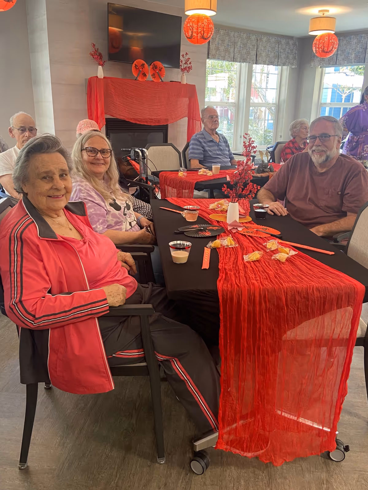 A group of elderly people sitting around a table decorated with a red table runner, red floral centerpieces, and snacks in a well-lit room with large windows. The room has a fireplace with red decorations and a TV mounted above it. The people appear to be enjoying a social gathering.