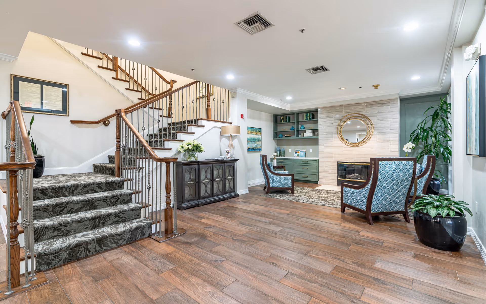A spacious and well-lit interior common area featuring a carpeted staircase with wooden handrails on the left, a dark wooden console table with a lamp and flower arrangement, two patterned armchairs facing a modern fireplace with a round mirror above it, built-in shelves with decorative items, and several potted plants. The floor is wood with a small area rug in front of the fireplace.