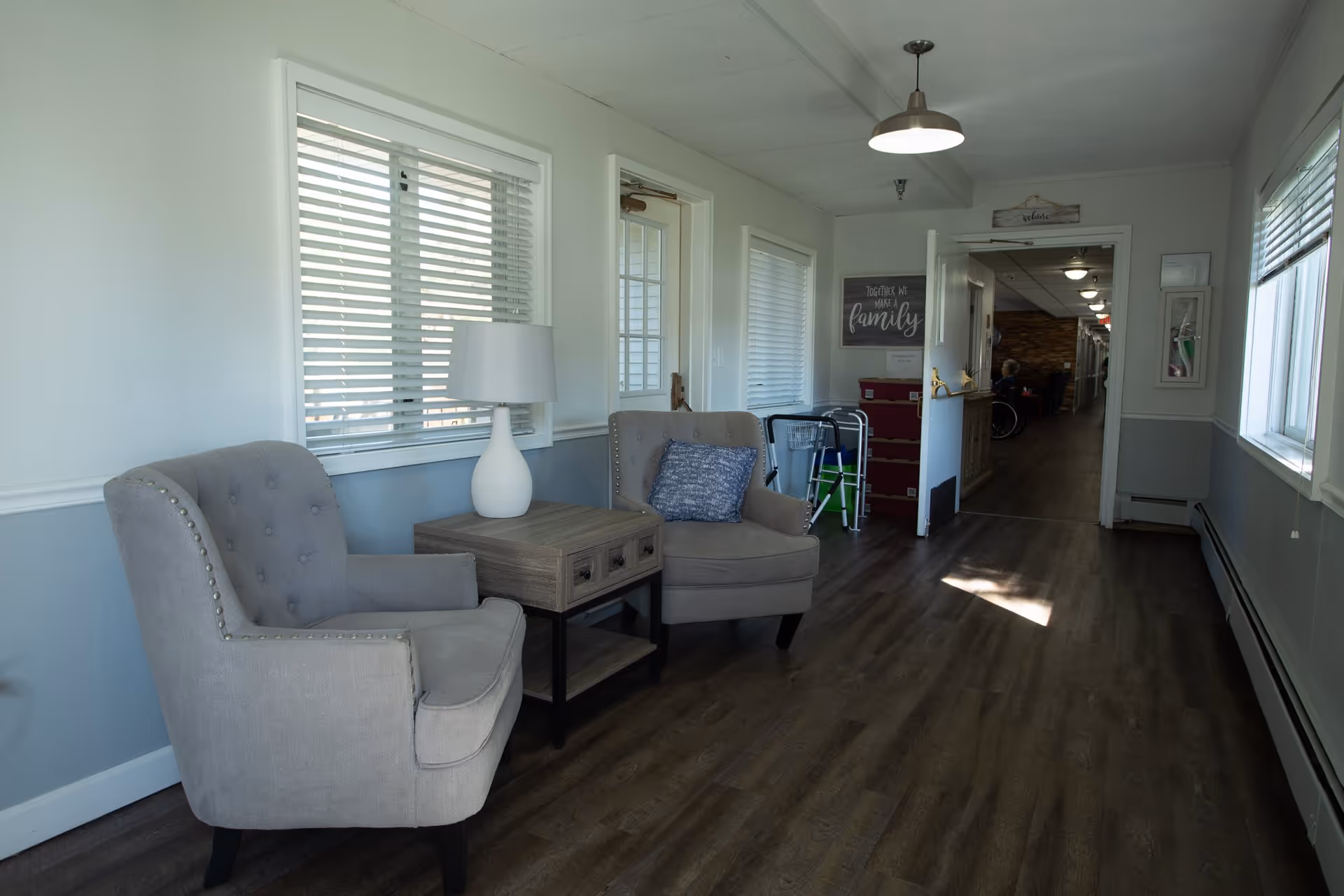 Hallway seating area with two upholstered chairs, a side table and lamp along a bright corridor in a senior living facility.