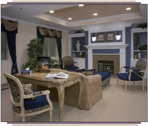 A cozy living room with a beige sofa, two upholstered armchairs with floral patterns, a wooden desk with an open book, and a fireplace with decorative items and framed pictures above it. The room has recessed ceiling lights and windows with dark blue curtains and gold valances.