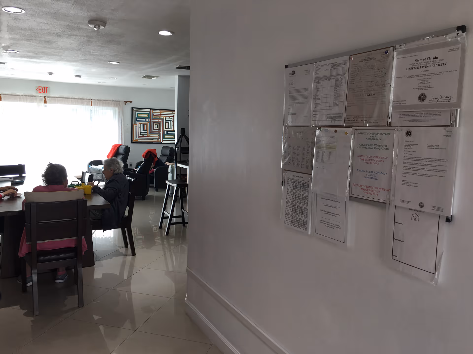 Interior view of a senior living facility showing a group of elderly people sitting around a table engaged in conversation. In the background, there are reclining chairs with red blankets draped over them and a colorful geometric wall art. On the right side, a bulletin board with various notices and documents is mounted on a white wall.