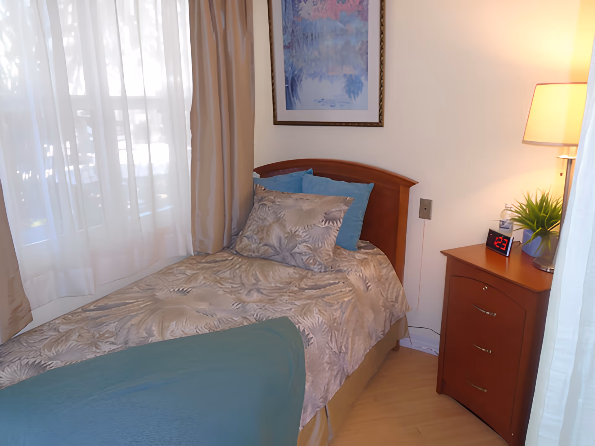 A cozy bedroom with a single bed covered in a patterned beige and blue bedspread, two blue pillows, and a wooden headboard. Next to the bed is a wooden nightstand with three drawers, a digital alarm clock displaying 1:23, a small green plant, a lamp, and a bottle of water. The room has light-colored walls, a large window with sheer white curtains and beige drapes, and a framed artwork hanging above the bed.