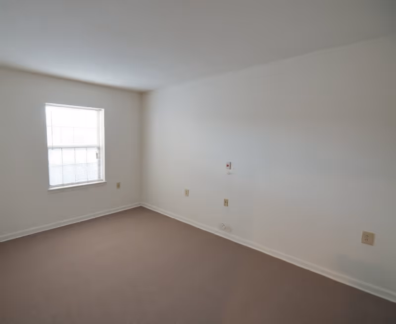 Empty room with beige carpet, white walls, a window with blinds on the left wall, and several electrical outlets on the walls.
