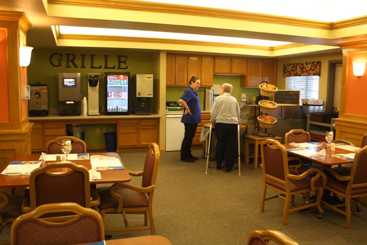 Dining area with wooden tables and chairs set with napkins and glasses. In the background, a beverage station with soda machines and a sign that reads 'GRILLE'. Two people are standing near the beverage station, one elderly person with a walker and another person in a blue shirt. The room has warm lighting and wood paneling.