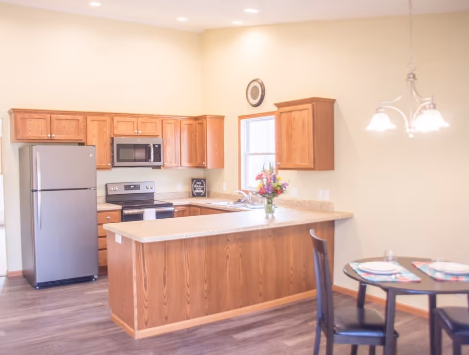 A bright kitchen with wooden cabinets, a stainless steel refrigerator, microwave, and stove. There is a countertop with a vase of colorful flowers and a small dining table set with two place settings and chairs. The walls are painted a light beige color and there is a window above the sink.