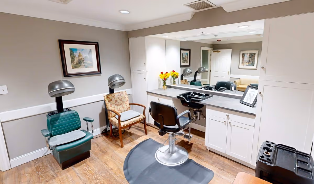 Interior view of a small salon area in a senior living facility with a green vintage hair dryer chair, a floral armchair, a black salon chair in front of a counter with a sink, a large mirror, and a vase with yellow flowers on the counter.