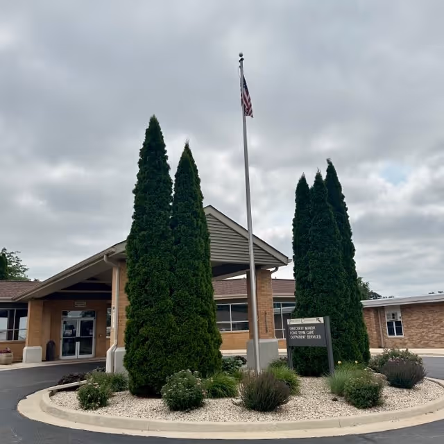 Exterior view of a single-story brick building with a covered entrance, surrounded by tall evergreen trees and landscaped bushes. An American flag is flying on a flagpole in front of the building under a cloudy sky.