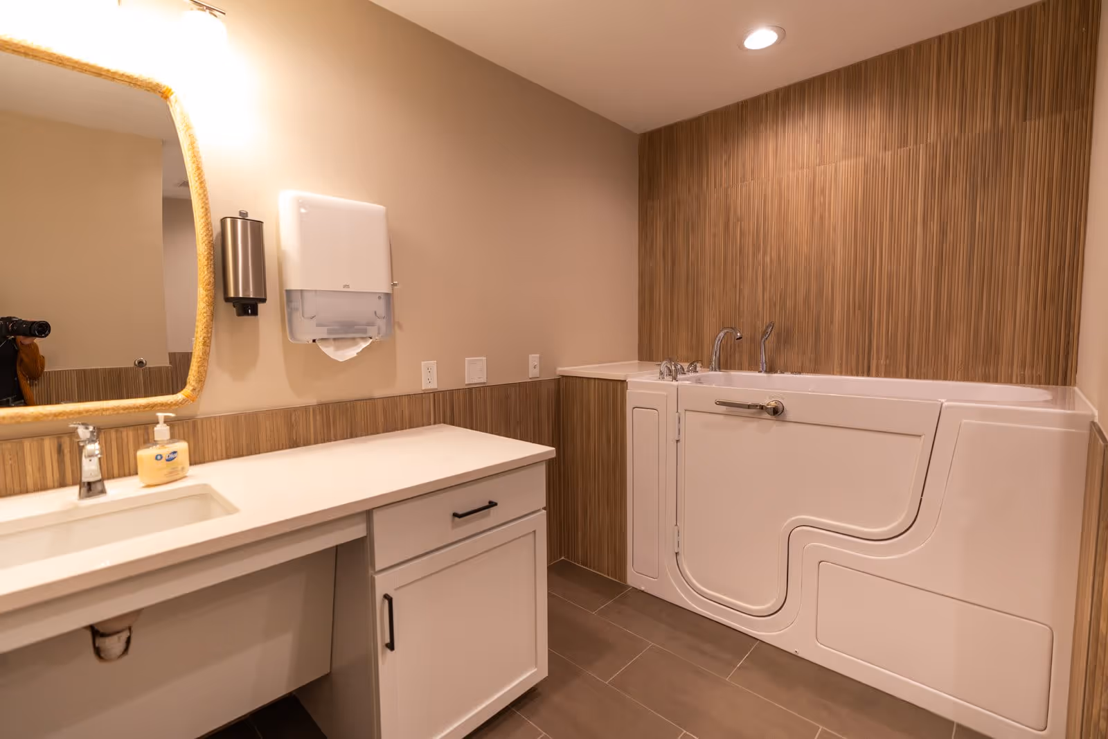 A bathroom featuring a white walk-in bathtub with a door and safety grab bar, a white countertop with a sink and faucet, a soap dispenser, a wall-mounted paper towel dispenser, and a mirror with a rope frame. The walls have a combination of beige paint and wood-textured tiles, and the floor is tiled in a neutral color.