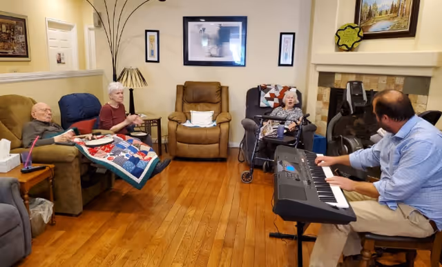 A group of elderly people sitting in a living room with wooden floors and beige walls. One elderly woman is clapping, another is sitting in a wheelchair with a colorful quilt, and an elderly man is seated on a recliner. A man is playing an electronic keyboard near a fireplace. The room is decorated with framed pictures and a tall floor lamp.