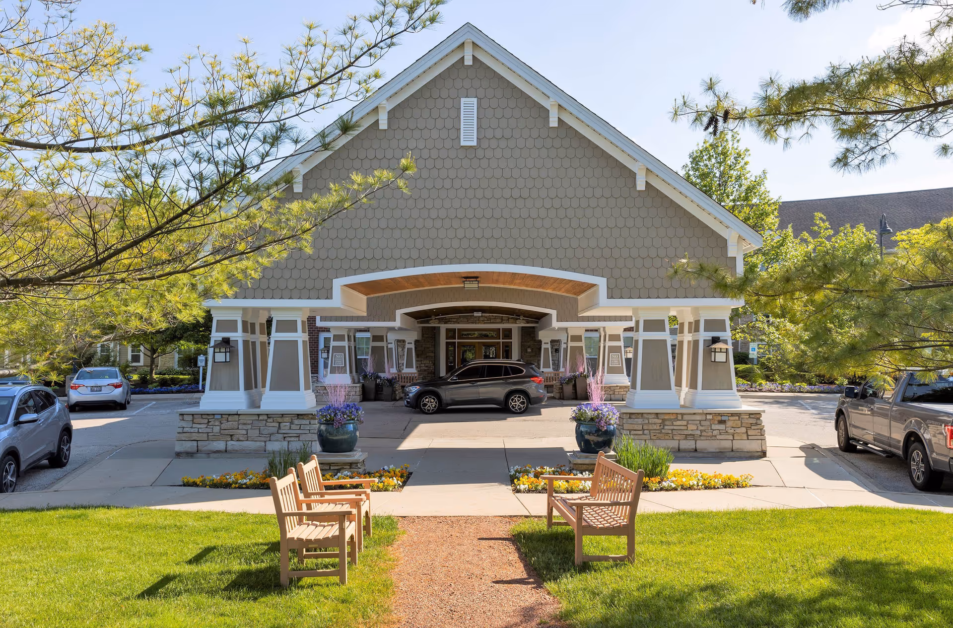 Front exterior view of a senior living facility named Chestnut Square at The Glen, featuring a covered entrance with a peaked roof, two large planters with purple flowers, parked cars on either side, and wooden benches on a grassy area in the foreground.