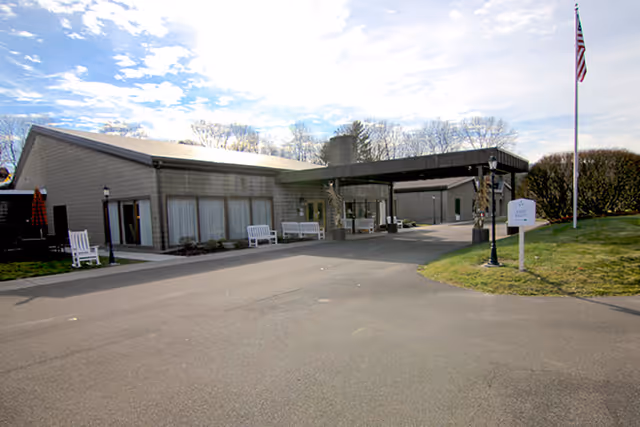 Single-story senior living facility entrance with a covered drop-off, benches, and an American flag on the lawn.