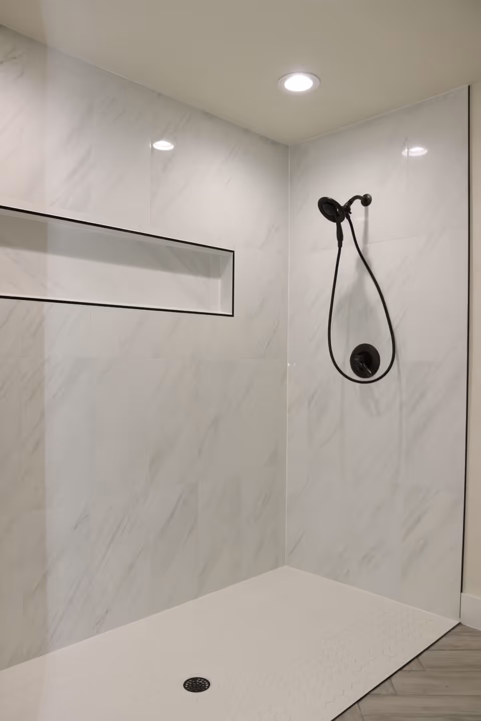 A modern walk-in shower with white marble-patterned wall tiles and a black handheld showerhead mounted on the wall. The shower floor has small white hexagonal tiles and a central drain. There is a recessed shelf built into the wall for storage.