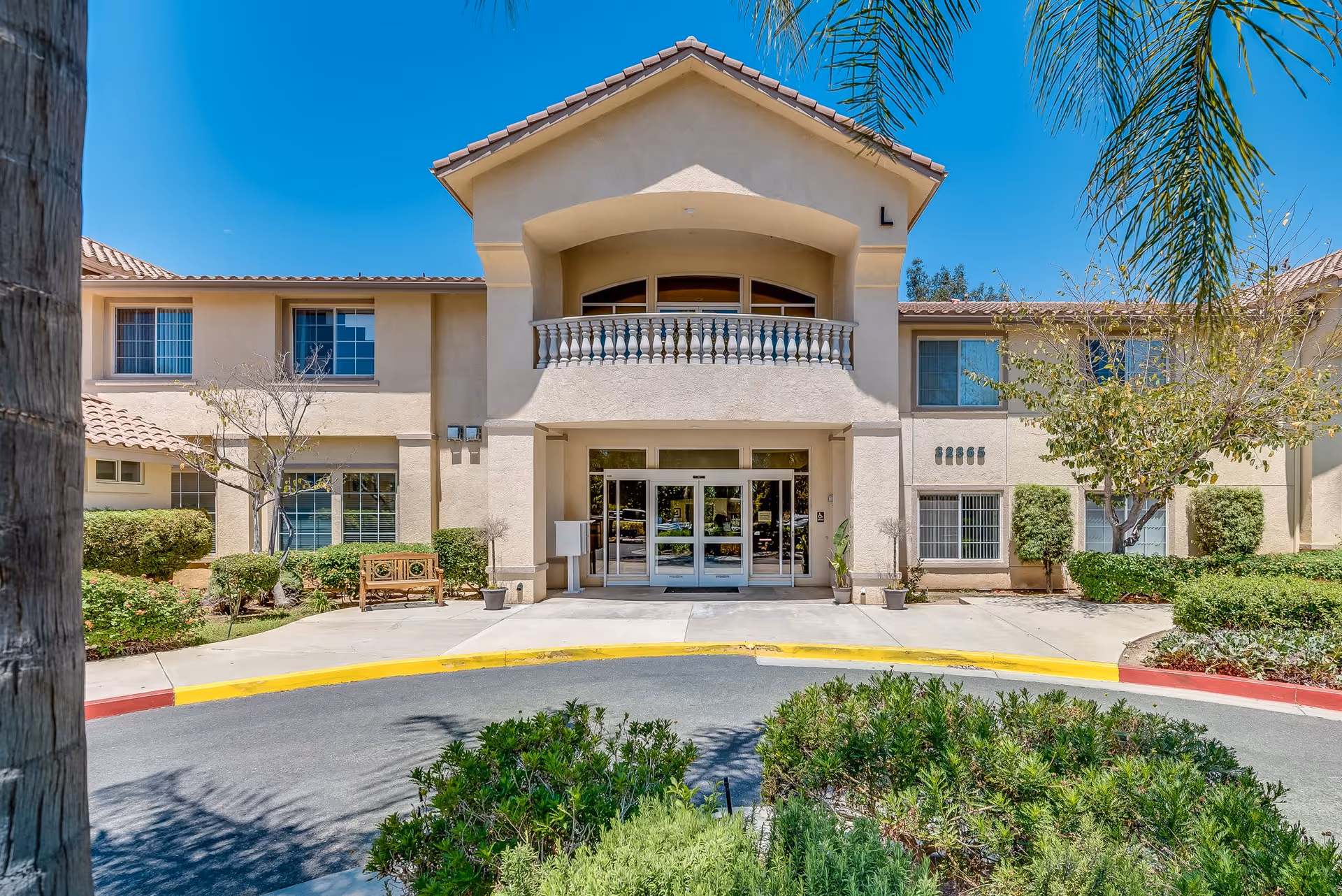 Front exterior view of Wildomar Senior Assisted Living facility with a beige stucco building, tiled roof, glass entrance doors, landscaped bushes, trees, and a bench near the entrance under a clear blue sky.