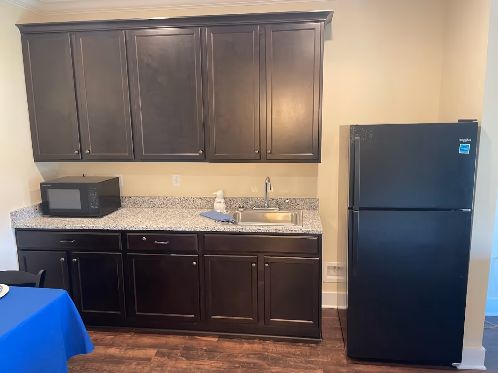A kitchen area with dark wooden cabinets, a granite countertop, a stainless steel sink with a faucet, a black microwave on the counter, and a black Whirlpool refrigerator. The floor is wooden, and a blue tablecloth is partially visible on the left side.