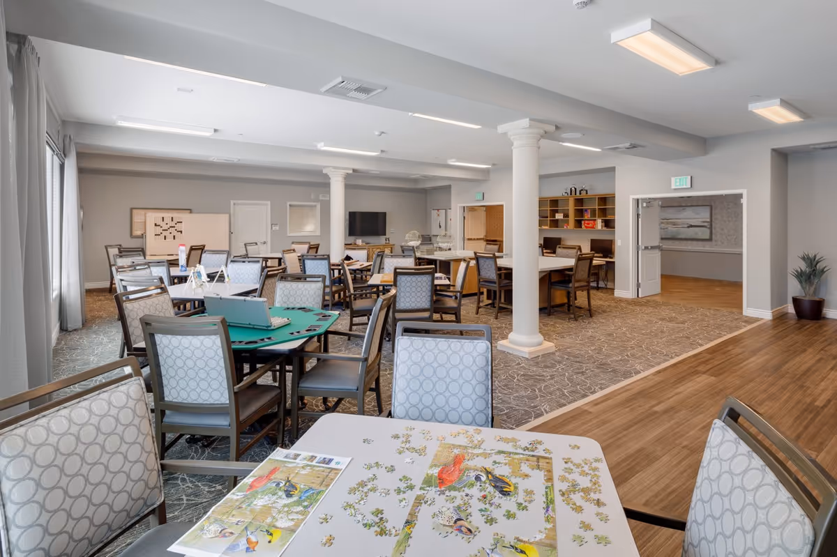 A spacious activity room in a senior living facility with multiple tables and chairs arranged for group activities. One table in the foreground has a partially completed bird-themed jigsaw puzzle and a reference image. Other tables have card games and board games set up. The room features neutral-colored walls, carpeted and wooden flooring, white columns, and overhead fluorescent lighting. There is a TV mounted on the far wall and shelves with books and supplies.